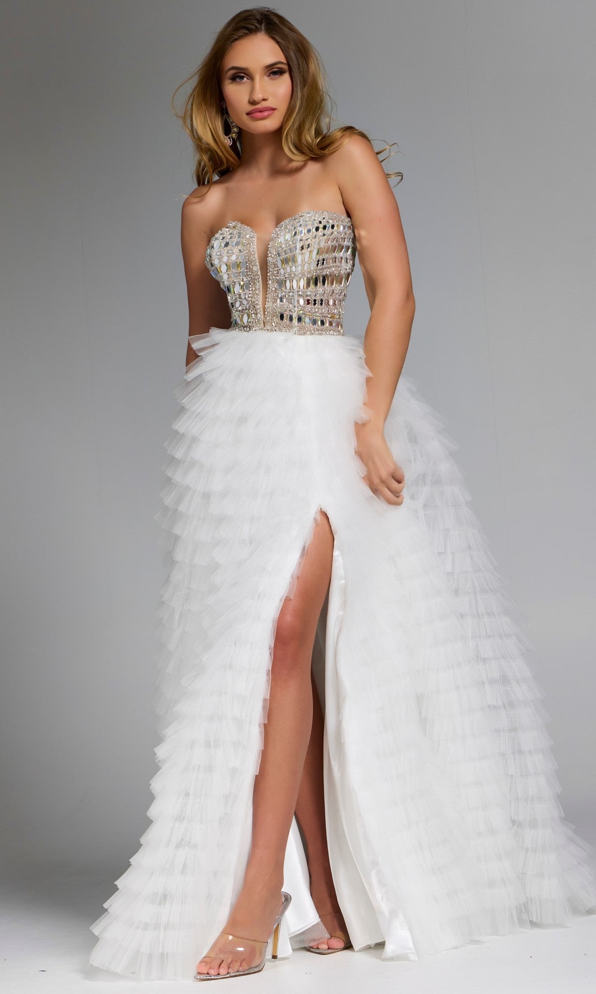 A woman stands against a plain background wearing the Long Prom Dress: Jovani 43412 in Off White, featuring a strapless beaded corset bodice, ruffle tulle skirt with a high front slit, and clear high-heeled sandals.